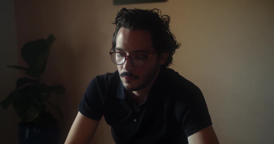 Man with glasses and a black polo shirt sits at a table and reads a book in a room