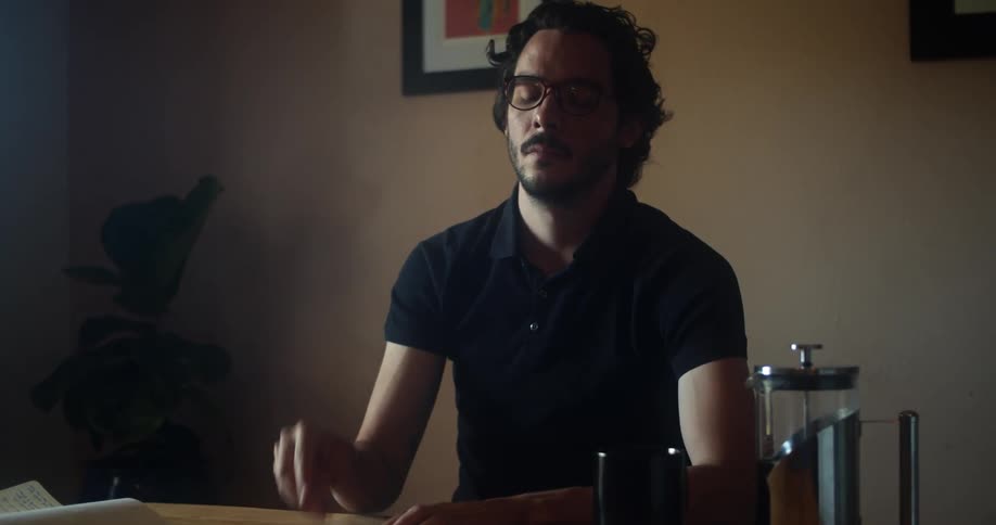 A young man with a beard puts down his book and sighs as he takes off his glasses