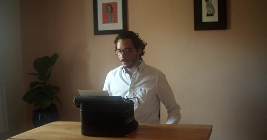 A man with glasses sitting on a chair in front of a table with a typewriter