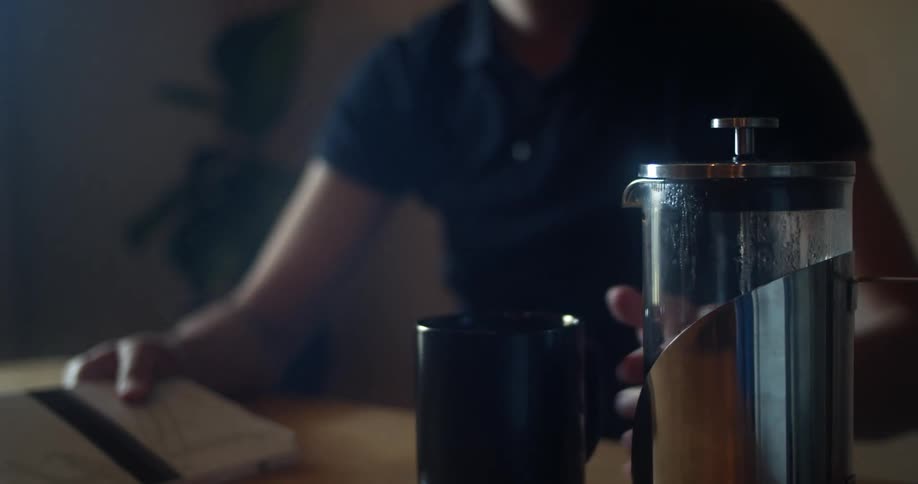 A man reading a book at a table pours coffee from a thermos into his mug and takes a drink before returning to his reading