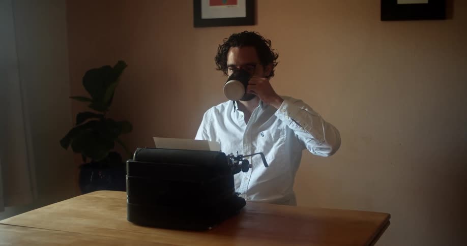 A man sitting at a mechanical typewriter reaches for and drinks from a large mug