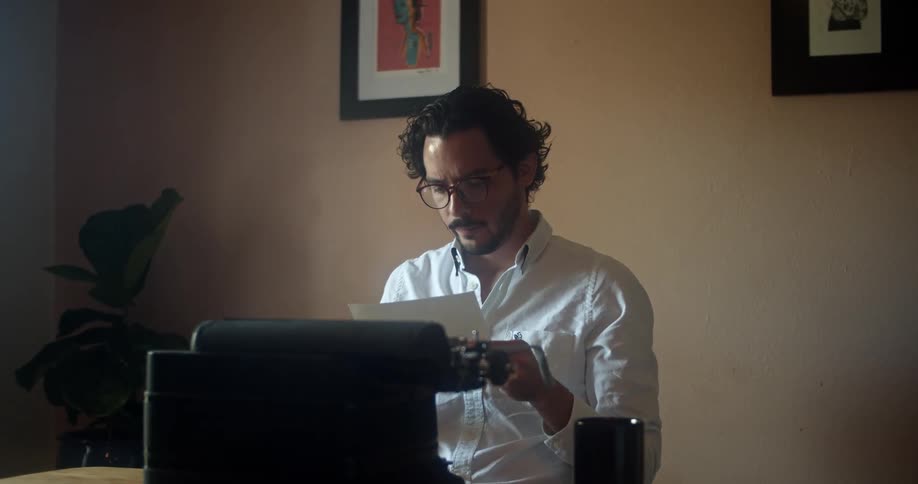 A man with glasses sitting at a desk taking a paper from a typewriter