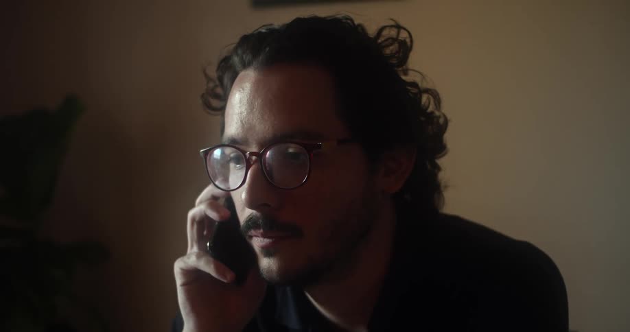 A man sitting indoors talks on a smartphone before moving it away from his face to look at the screen
