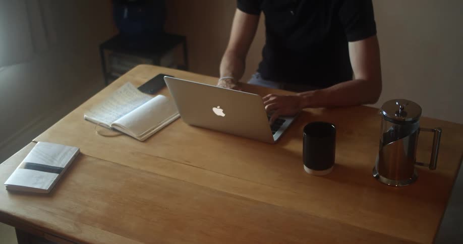 Man with a black polo shirt sits at a table and uses a macbook laptop