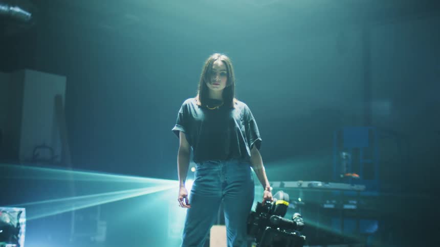 A woman stands in a dark film studio holding a large lamp while surrounded by electronic equipment