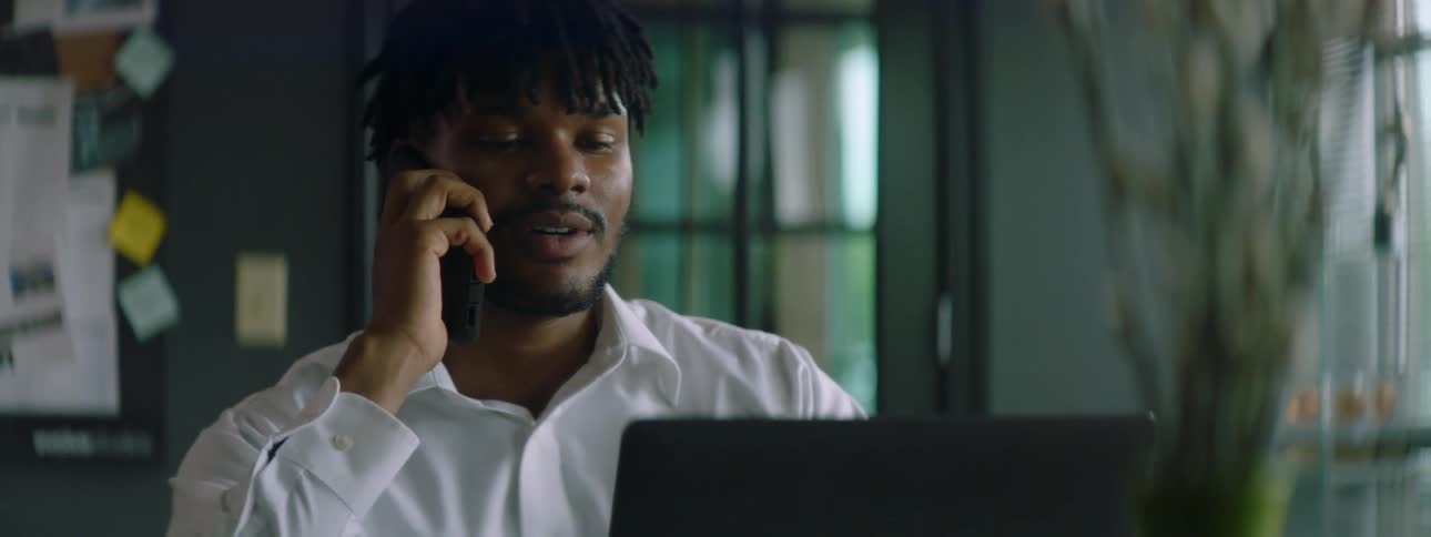 A young man typing on a laptop at a table lifts his smartphone and answers a call