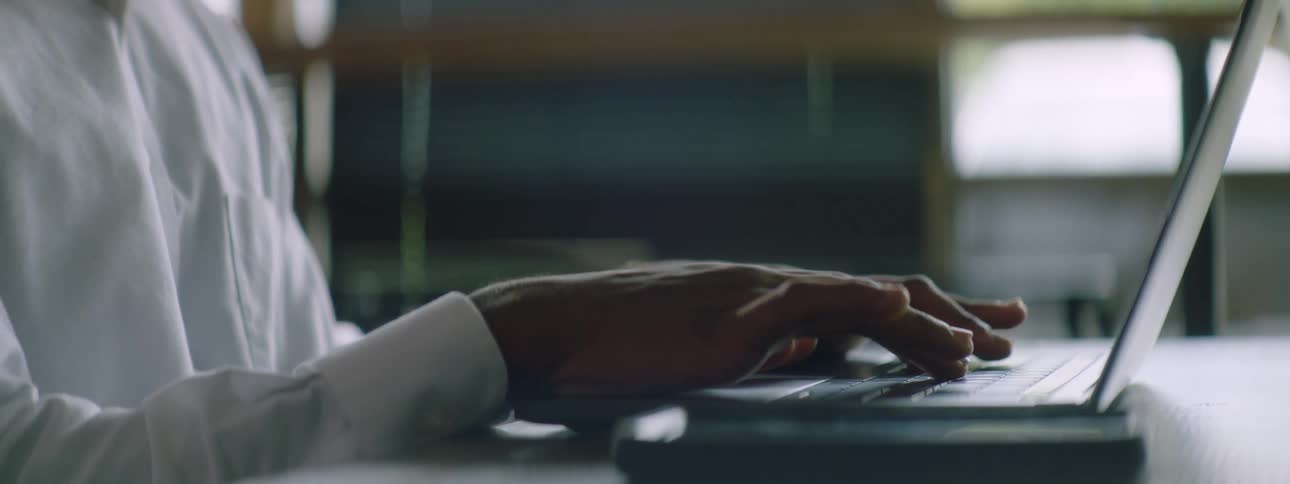 Black man with braided hair a beard and a white shirt sits at a table and uses a laptop in a room