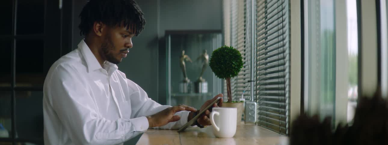 Black man with braids a beard and a white shirt sits at a table and uses a grey tablet