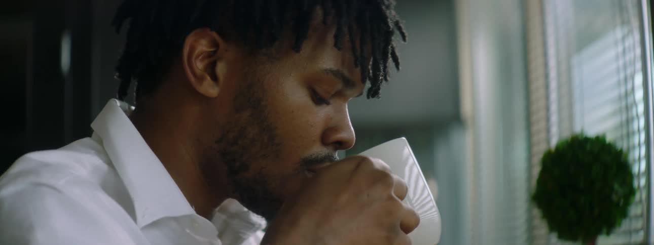 A young man sits at a desk at home and scrolls through his phone while drinking coffee