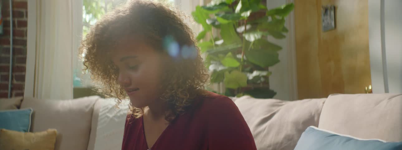 Woman with red curly hair and a shirt sits on a sofa and uses a grey laptop in the living room