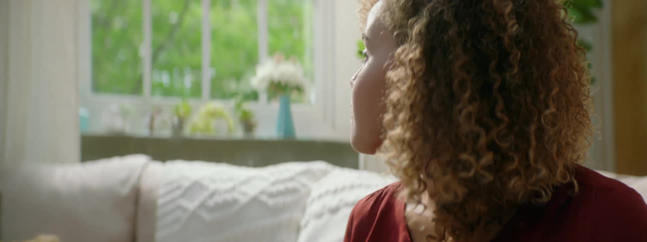 A young woman sitting on a sofa in a living room while typing raises her head to gaze out the window before returning to her typing