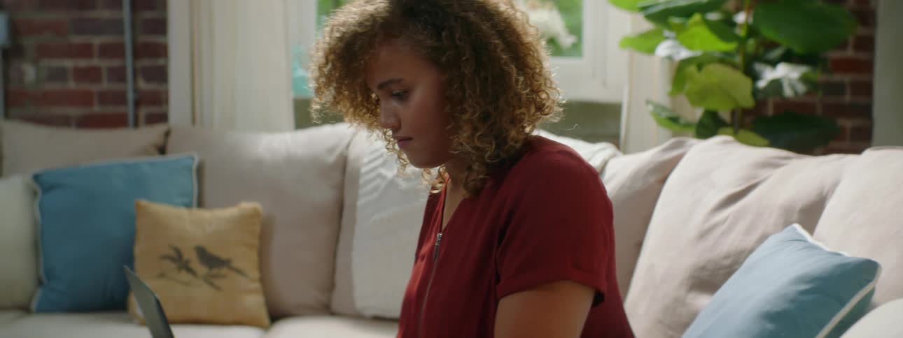 Woman with red curly hair black pants and a red blouse sits on a beige sofa and opens her laptop in the living room