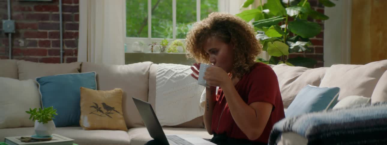 A woman sitting on a couch typing on a laptop and sipping on tea