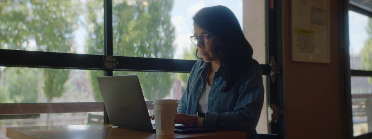 Brunette woman with wavy hair glasses and a blue shirt sits at a table and uses a grey laptop in a cafe