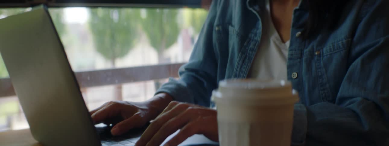 A young woman sitting at a table near a window types on her laptop while talking on a smartphone behind held to her face by her shoulder