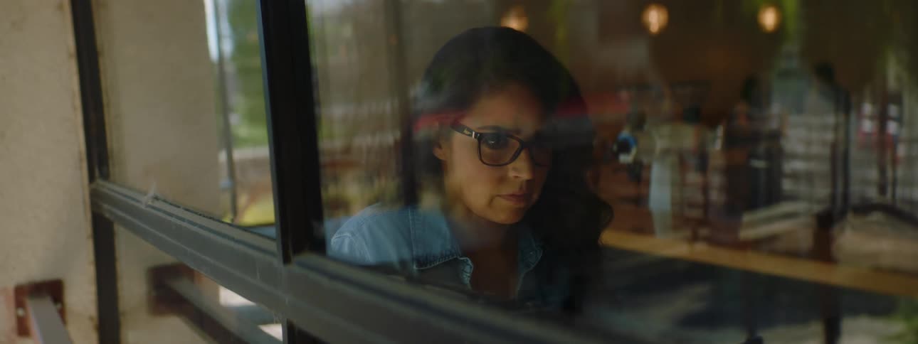 Woman with glasses and a blue shirt sitting at a table and using a laptop seen from outside of a cafe