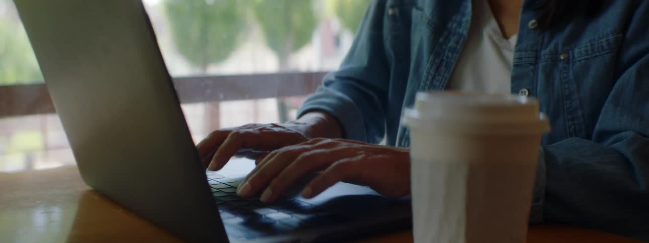 Woman with wavy hair a blue shirt and glasses types on a laptop while talking on a smartphone in a cafe