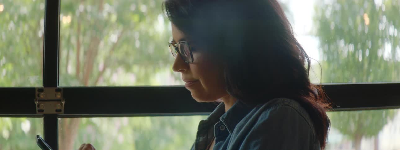 Young woman wearing glasses sitting on a table checking her smartphone next to a laptop