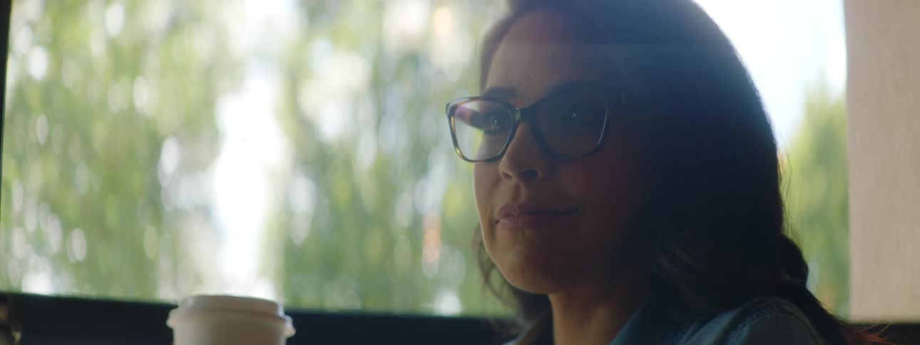 Brunette woman with wavy hair and glasses drinks coffee and smiles while using a laptop in a cafe