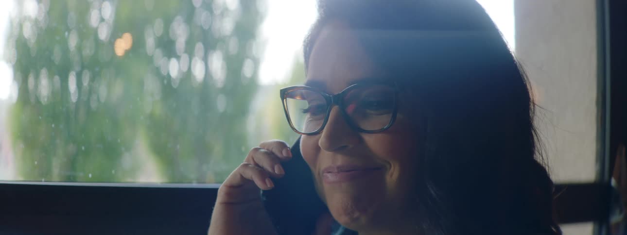 A woman laughs as she talks on the phone while sitting in front of a large window