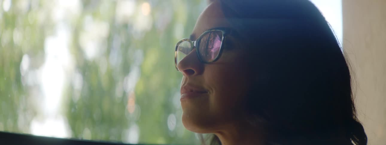 A woman sitting indoors by large windows is typing on a laptop and turns to smile at the camera