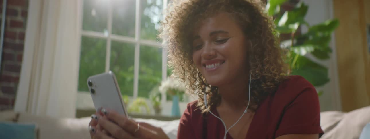 Woman with red curly hair and a red blouse sits on a sofa makes a video call on a smartphone with earphones in the living room at home