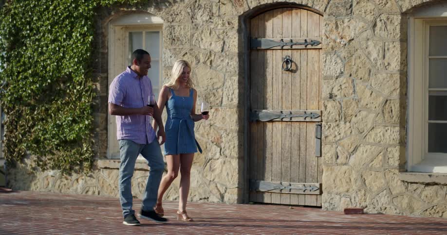 A man and a woman walking and talking while holding wine glasses with wine with a building in the background