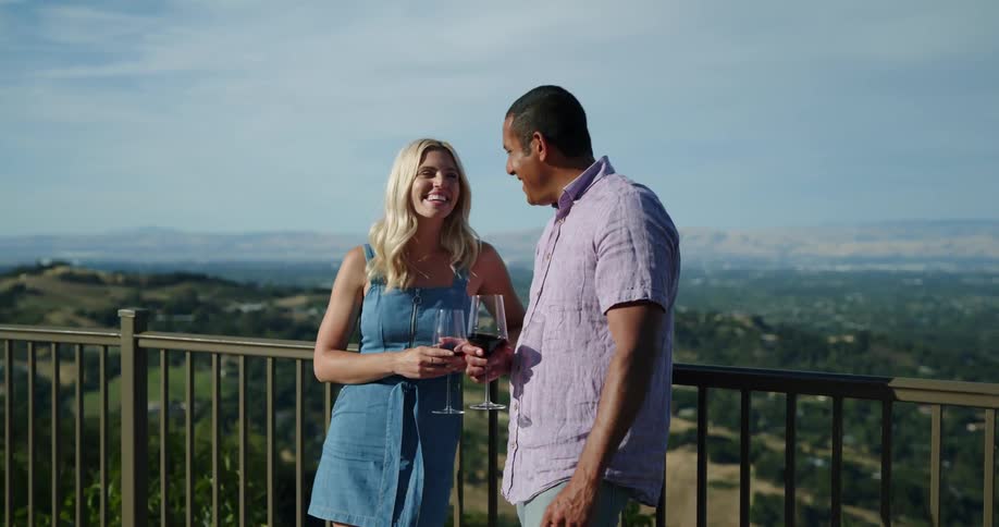 A man and woman stand on a high balcony holding wine glasses