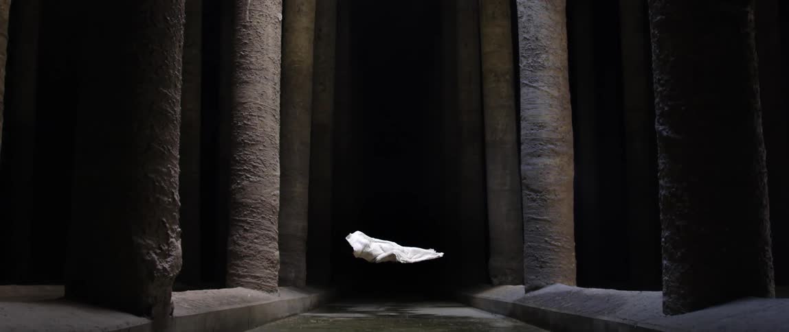 White fabric shape floats in mid air in stone floored corridor bordered by tall columns