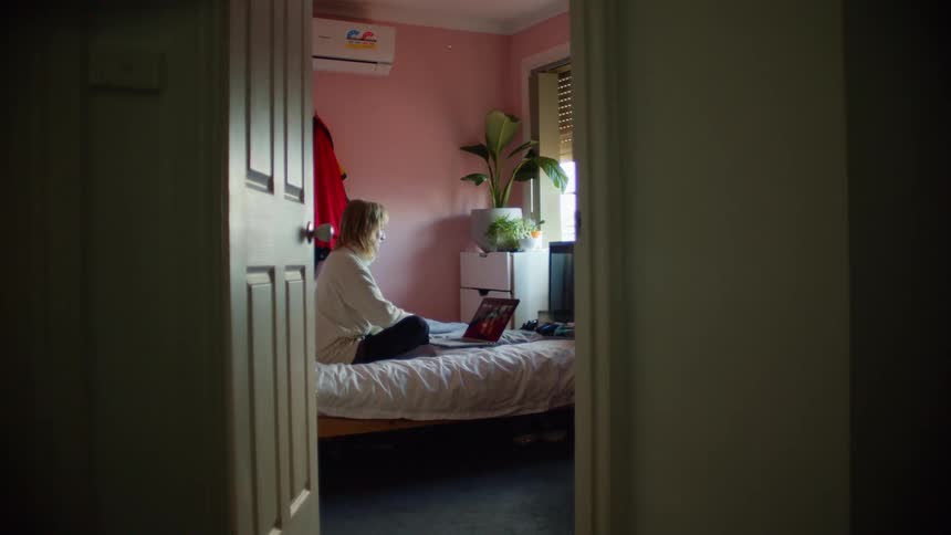 A woman sitting cross legged on her bed uses her laptop to participate in a video chat