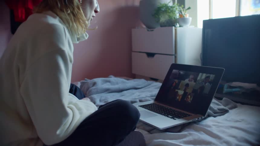 A young woman sits on her bed and video chats with friends on a laptop