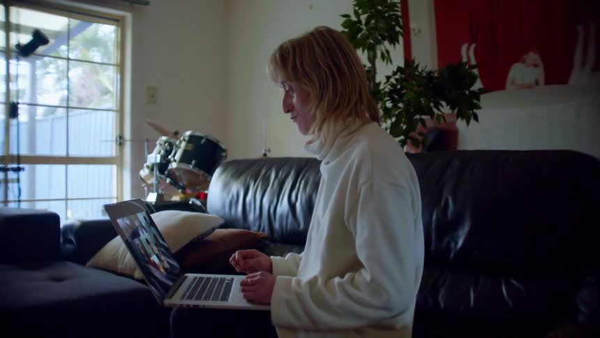Person sits on couch in living room containing drum set talking to friends via laptop