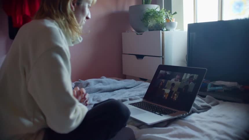 A woman uses a laptop to participate in a video chat while sitting on her bed in her bedroom