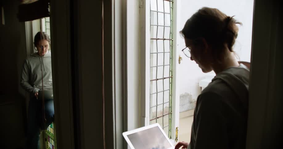 A woman sits in a doorway near a mirror and types on her laptop