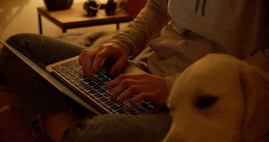 A woman sits on a couch with a laptop and her dog cuddles up beside her