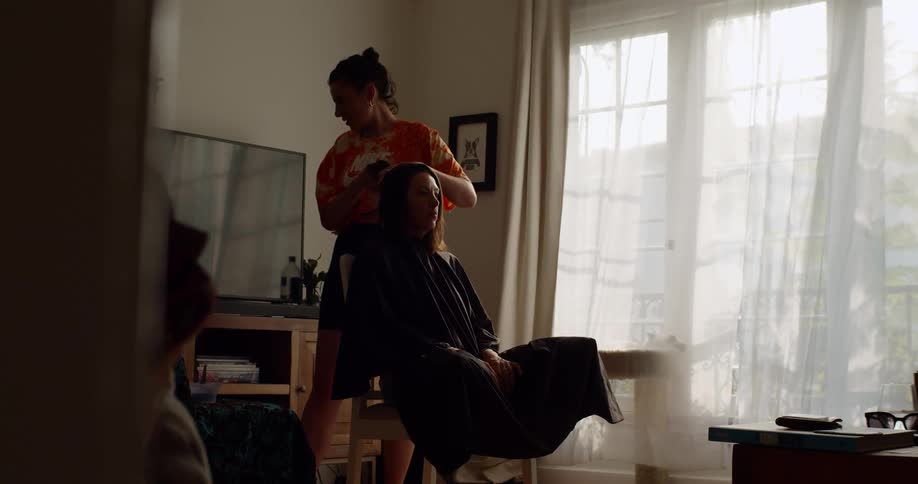 A woman stands behind a seated woman and holds her hair up then reaches behind for comb