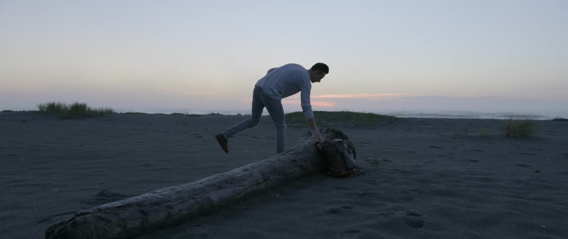 A man walking on a beach takes off his backpack and sits on a tree trunk to look at the sunset