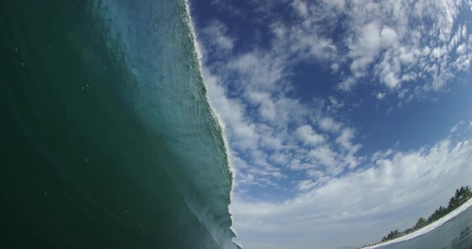 A surfer lying prone on their surfboard is swept up and over a large wave as it rolls towards the beach