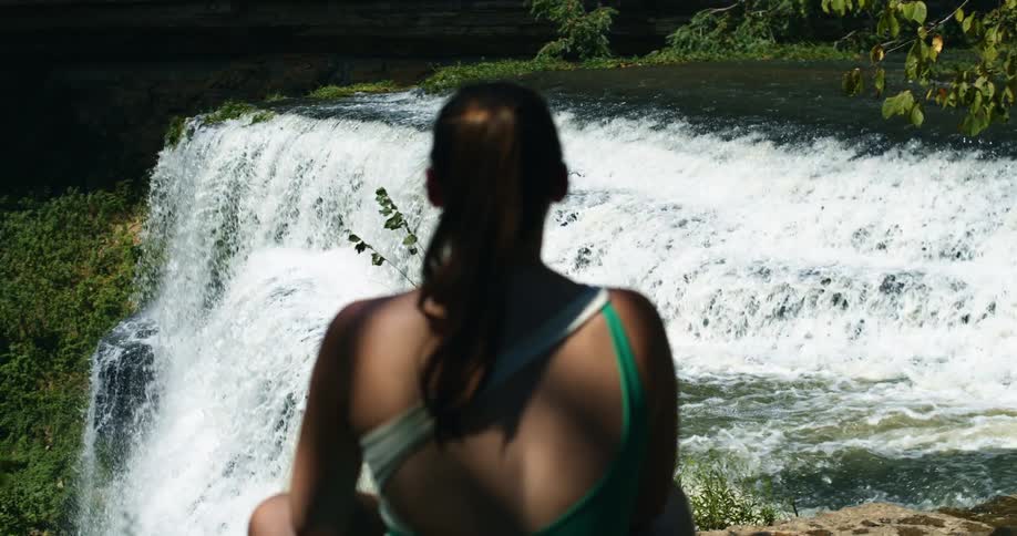 A teenage girl sits on a cliff and watches a waterfall crashing in to the river below