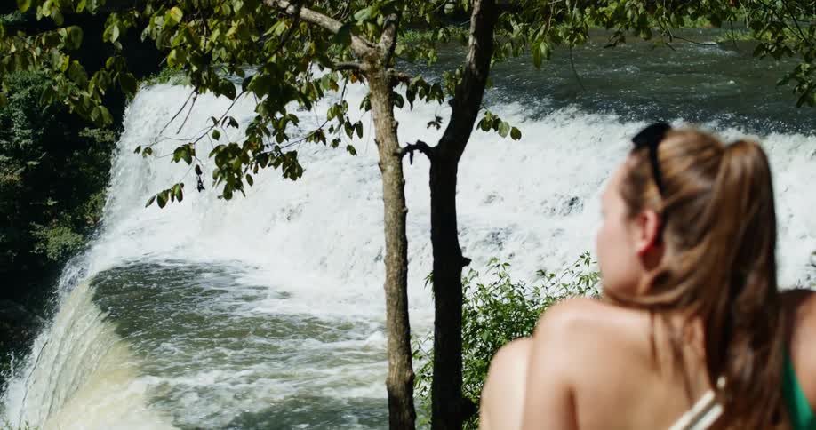 A woman with sunglasses sitting down with a river and a waterfall in the background