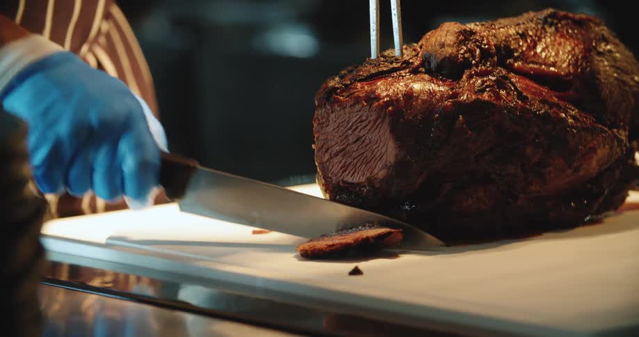 A man in a gloves and an apron holds a slab of beef still with a carving fork as he carves it with a knife