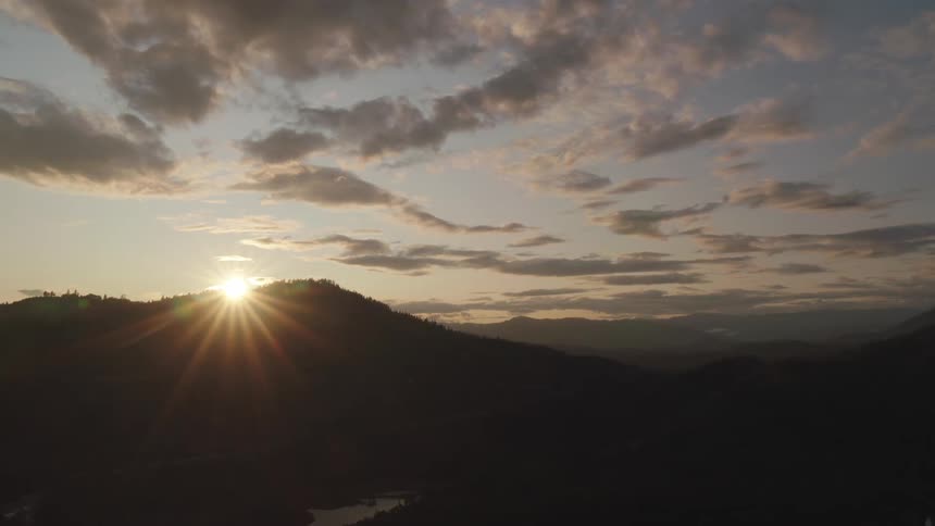 Sun sinks behind forested mountain bordering river under cloudy sky