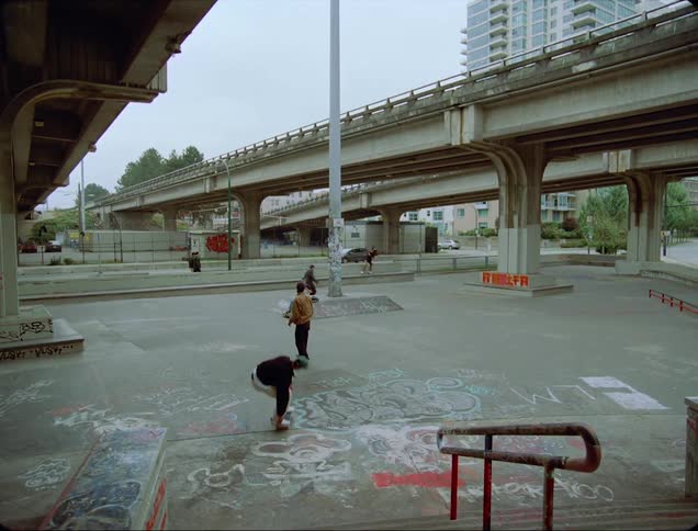 A group of skateboarders do tricks under a freeway bridge in a metropolis during the day