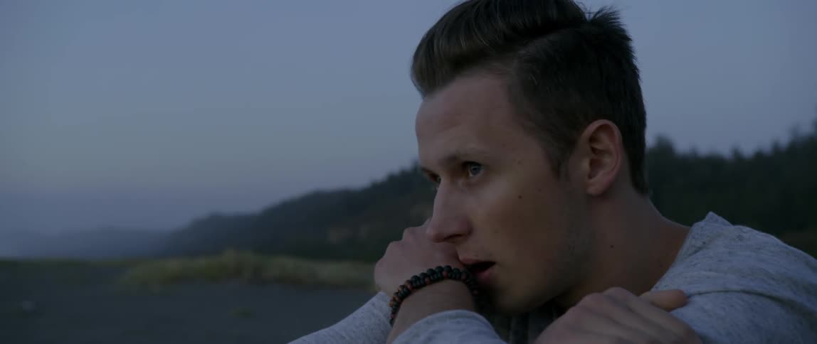 A man who seems slightly upset is sitting in a natural setting wrapping his arms around himself