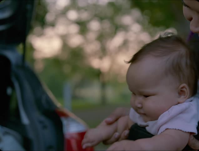 A woman waves the hand of a baby being carried in a baby carrier across her chest as another person waves at the child