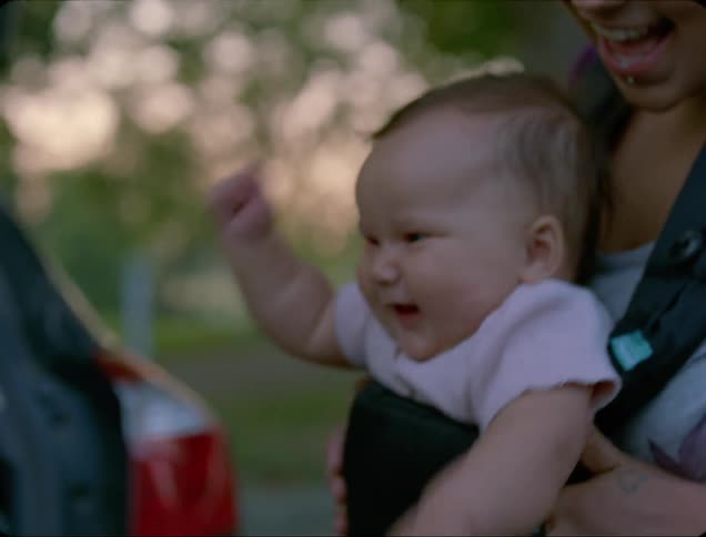 A woman carrying a baby in a baby carrier on her chest watches as the baby playfully swings her arms