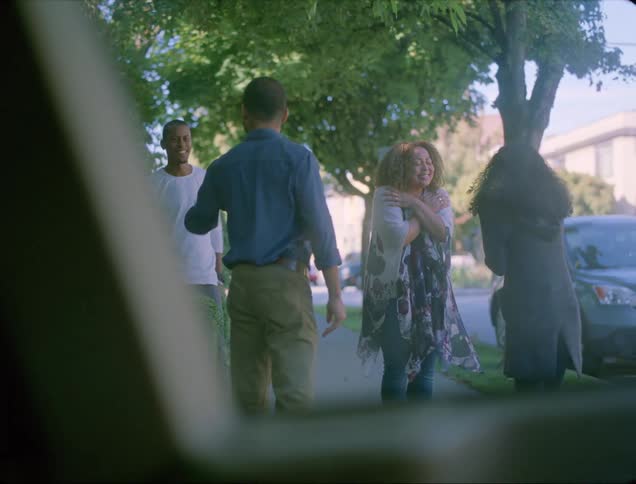 A man and a woman walking down stairs onto the pavement greeting another man and woman with trees and vehicles in the background