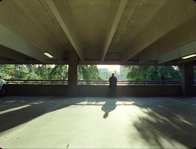 Man leans against rail in parking garage near car with city visible behind him