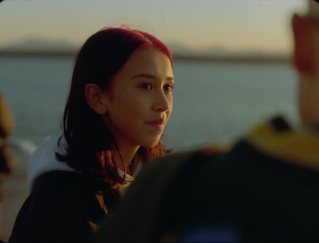 A young woman smiles while standing at the side of a lake alongside a man in the evening
