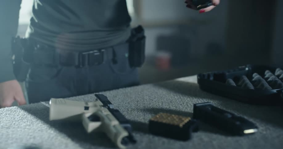 A woman takes an ammunition clip from a table covered in firearms and places it in a pouch on her belt